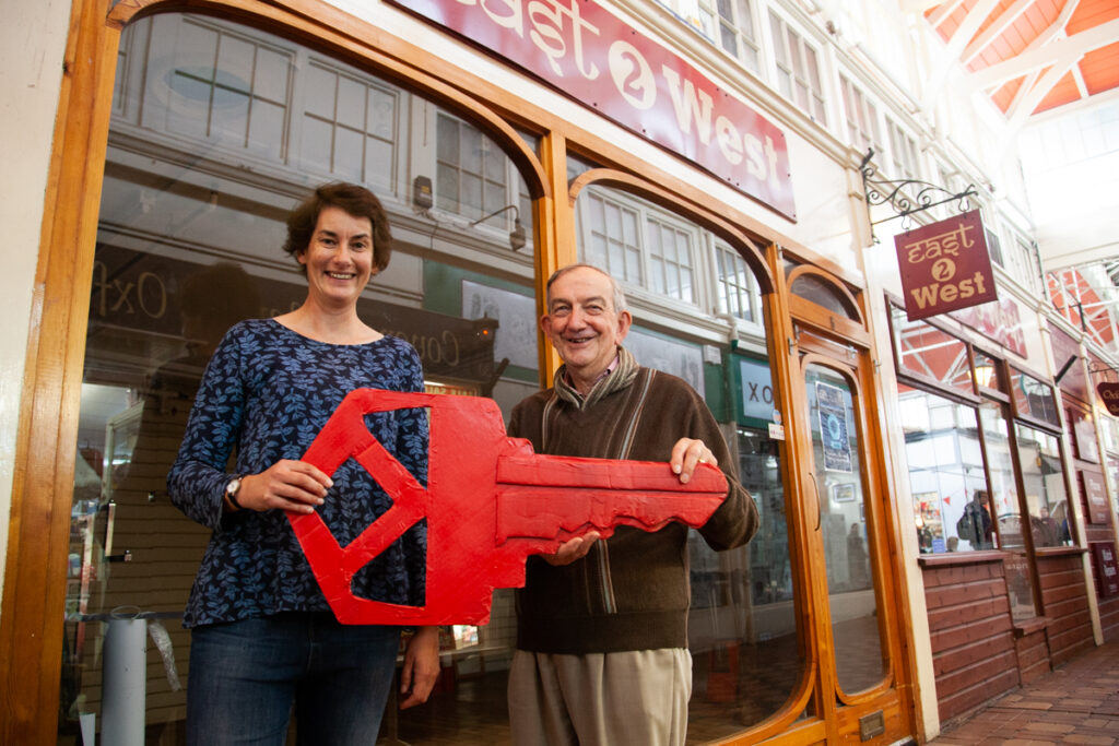 Two people hold a large red key outside and empty retail unit in Oxford's Covered Market