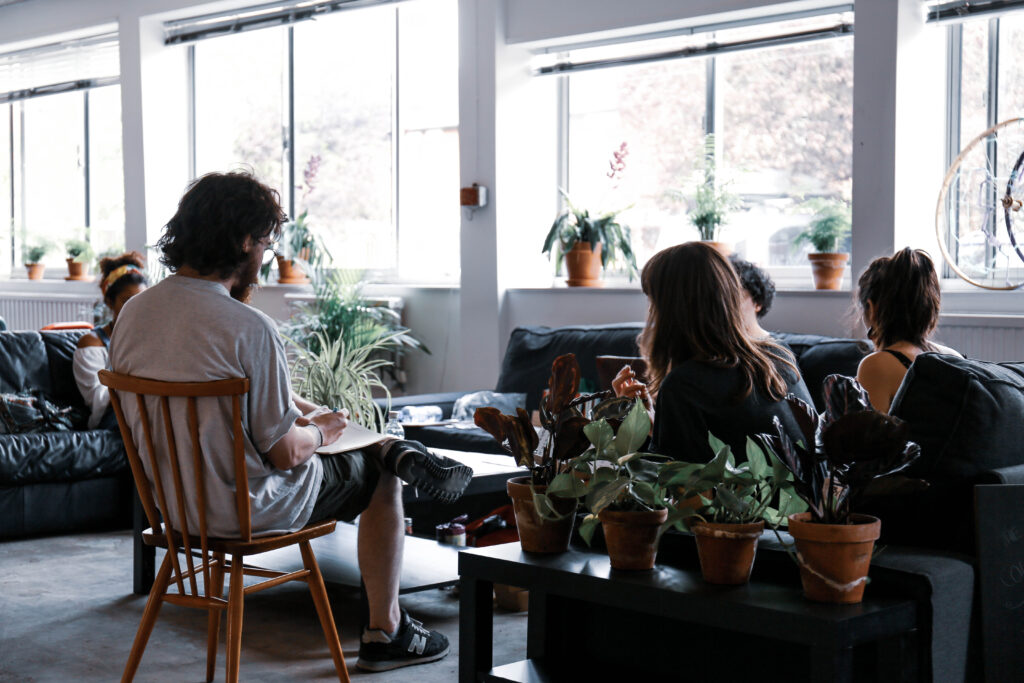 A picture of people sitting in a shared work space speaking with large windows behind them and lots of potted plants