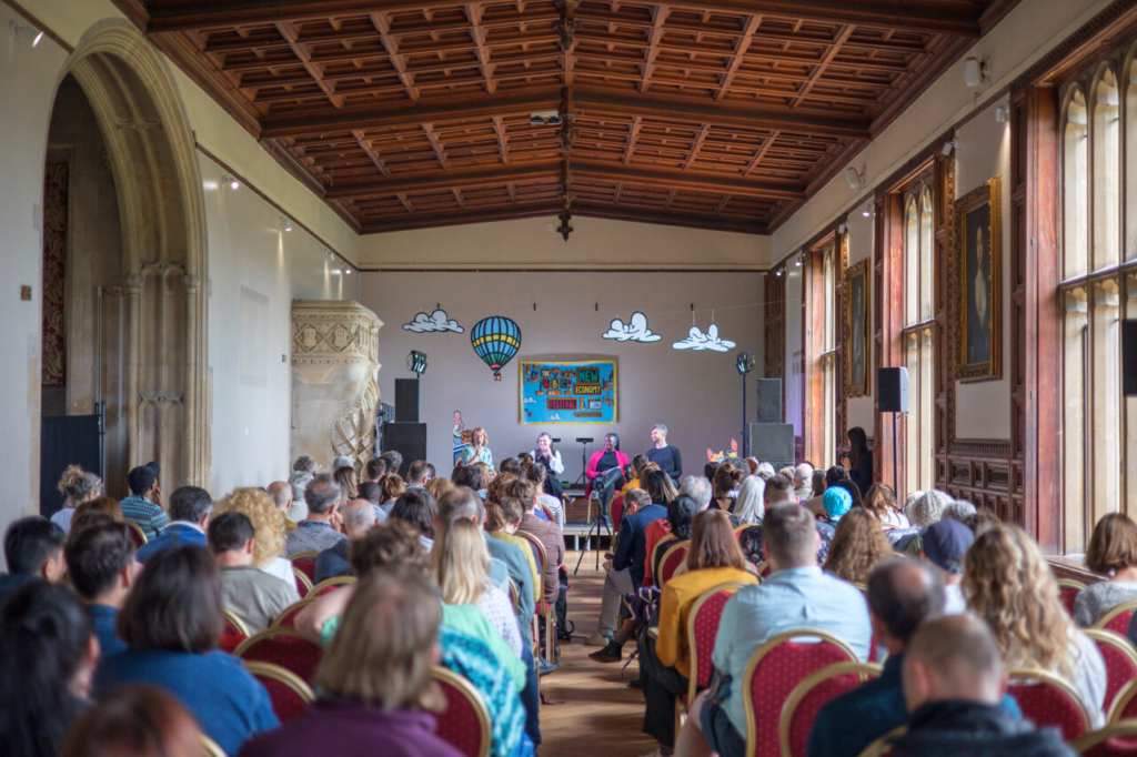 Wide format view of four people sat on stage at the end of a large hall with seated audience on either side