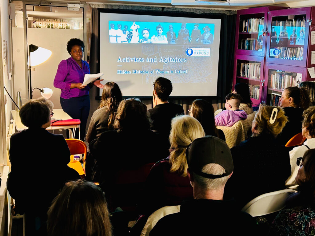 A woman sitting on a high stool in front of an audience, giving a talk. In the background, a screen reads "activists and agitators"