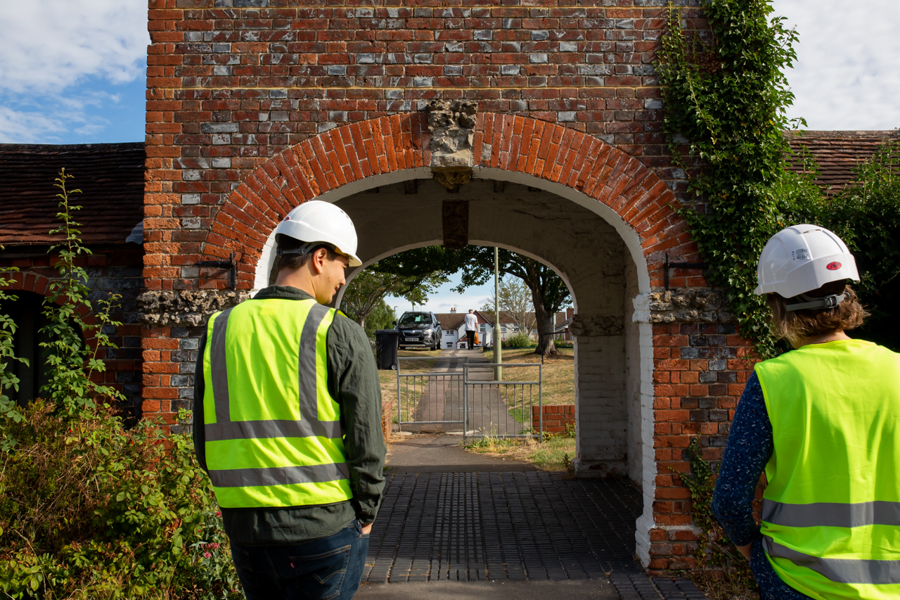 The Old Stables_small-9032 Two people in hard hats, viewed from behind, standing in front of a red brick structure