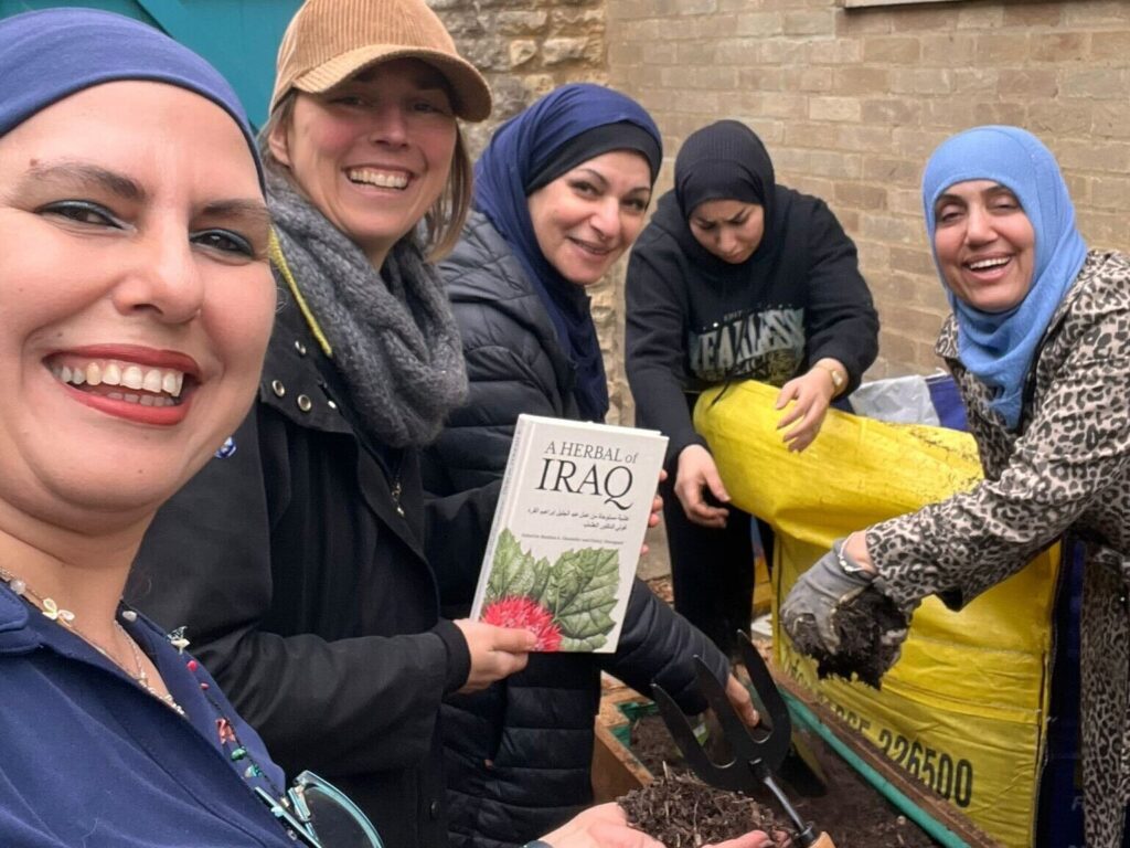 A group of women, filling a planter with compost, posing for a photo. One holds a book called "A herbal of Iraq"
