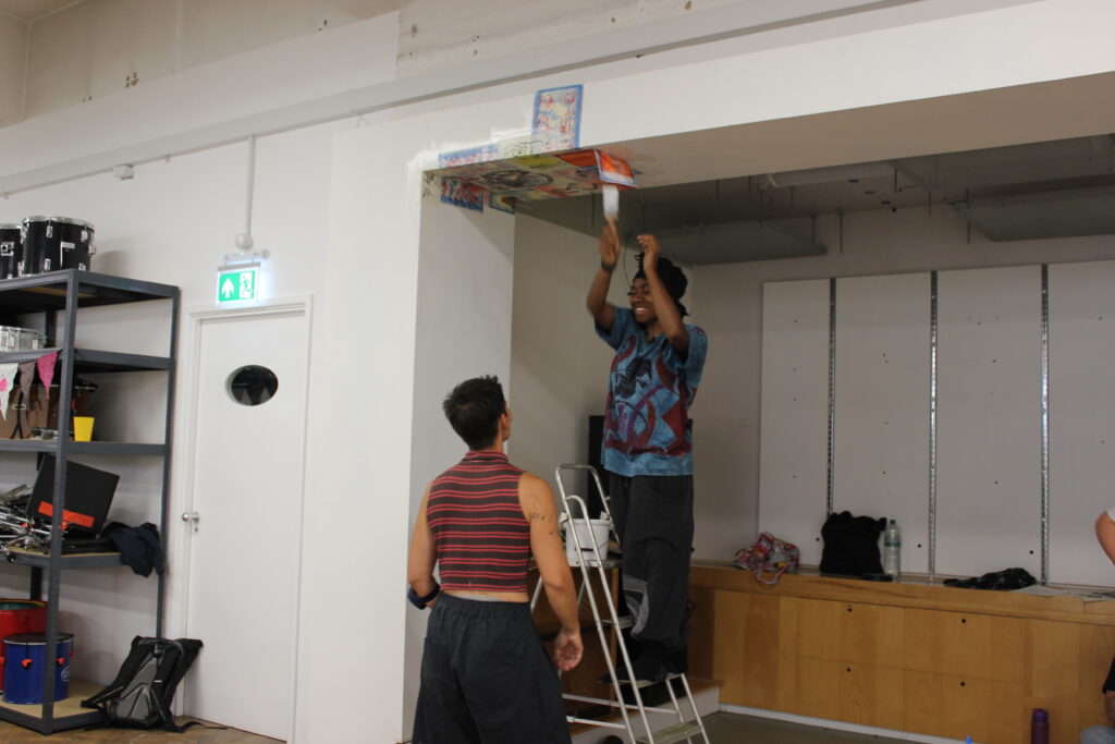 A young person standing on a step stool, laughing, pasting a flyer to a wall, while an adult looks on